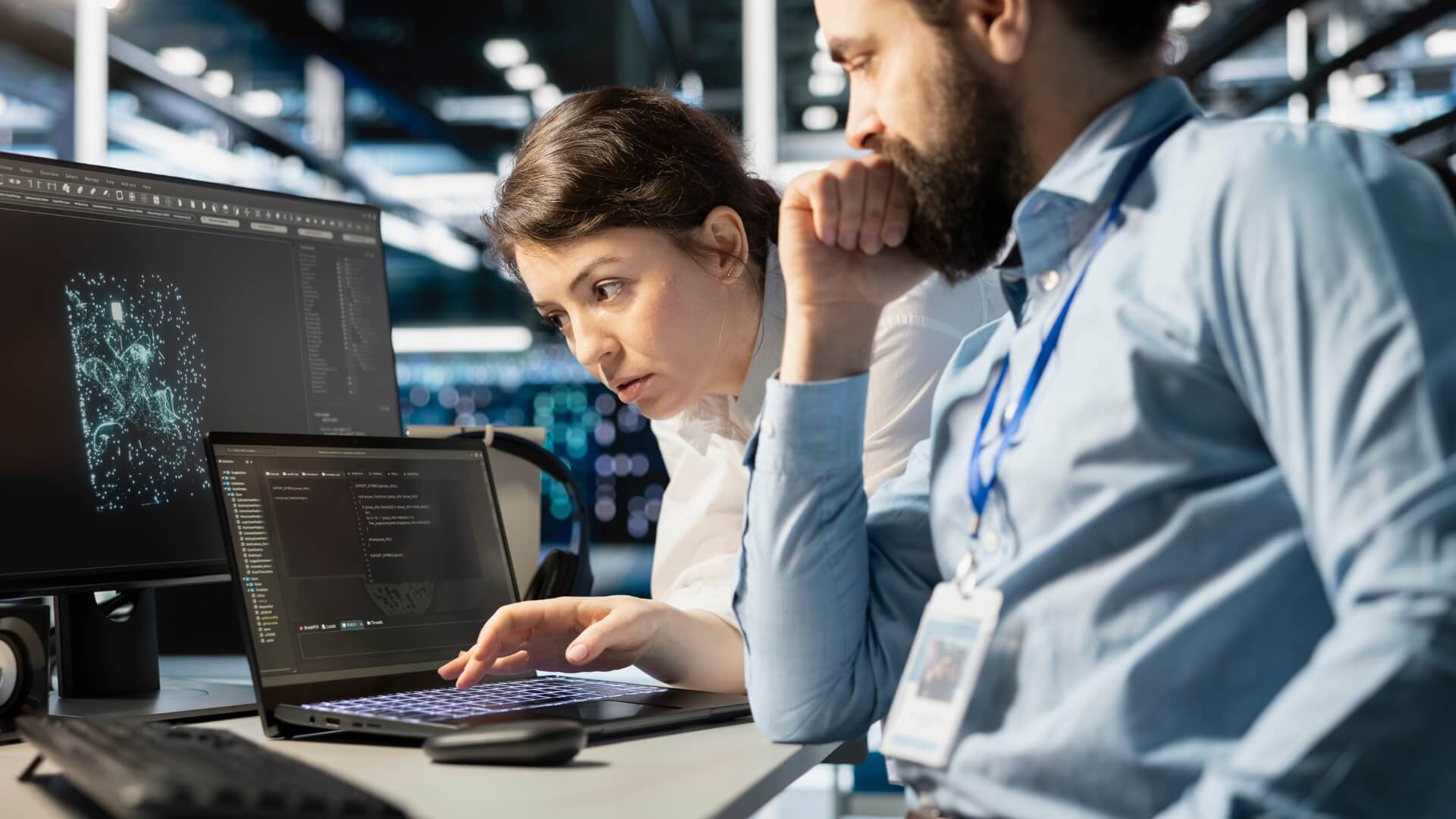 Two cybersecurity analysts collaborating at a workstation, reviewing code and network security data on multiple computer screens in a modern technology operations center. Professionals monitoring cybersecurity threats, software development, data analysis, and IT infrastructure management.