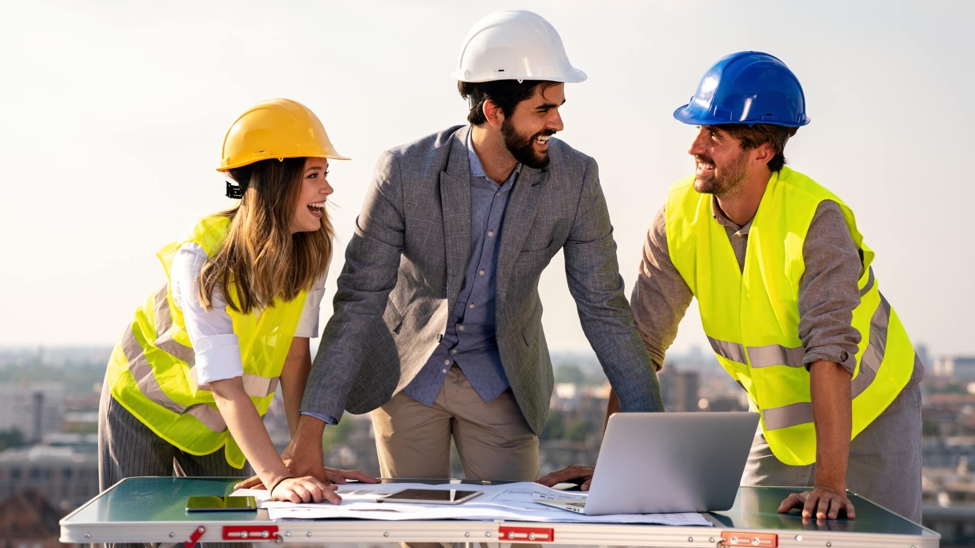 A commercial construction management team smiling and reviewing architectural blueprints on an outdoor rooftop site.