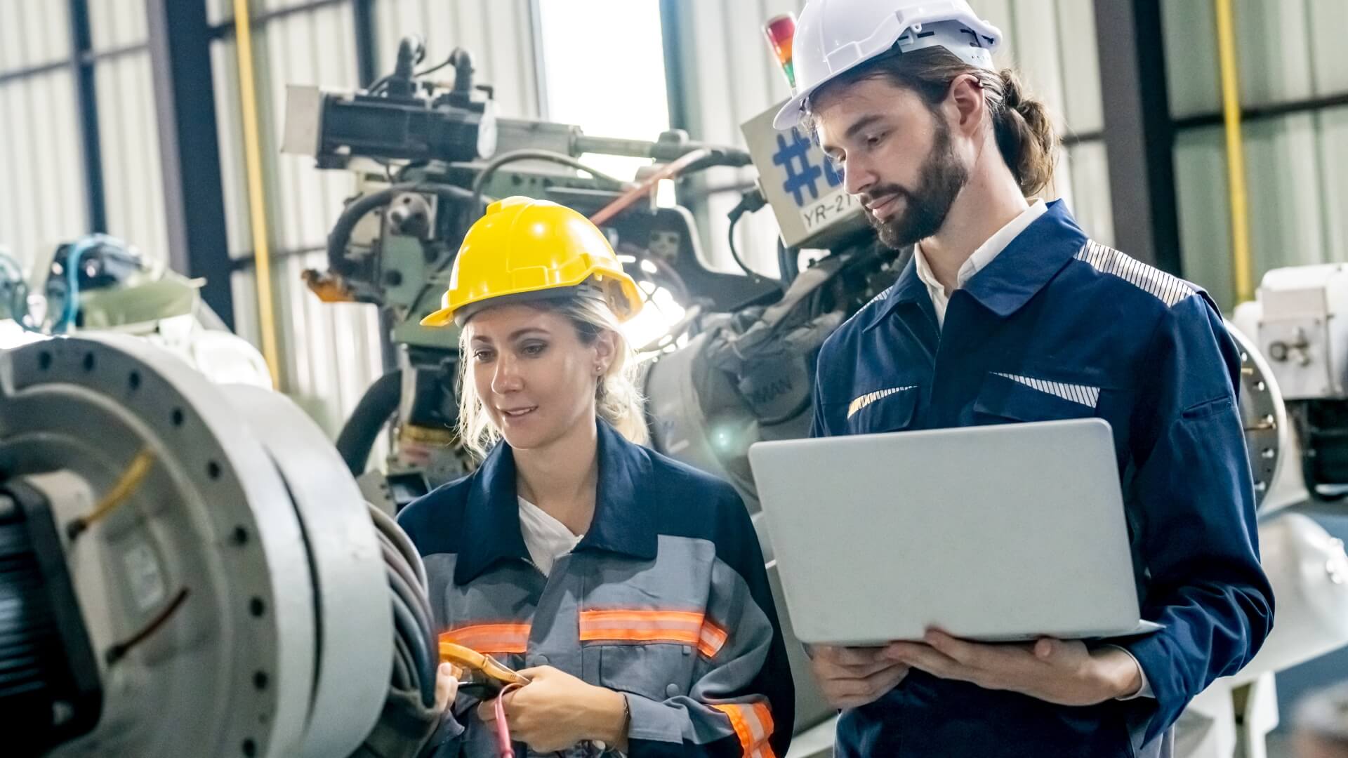 Two mechanical engineers wearing hard hats and safety gear using a laptop and digital multimeter to inspect heavy factory machinery on a manufacturing floor.