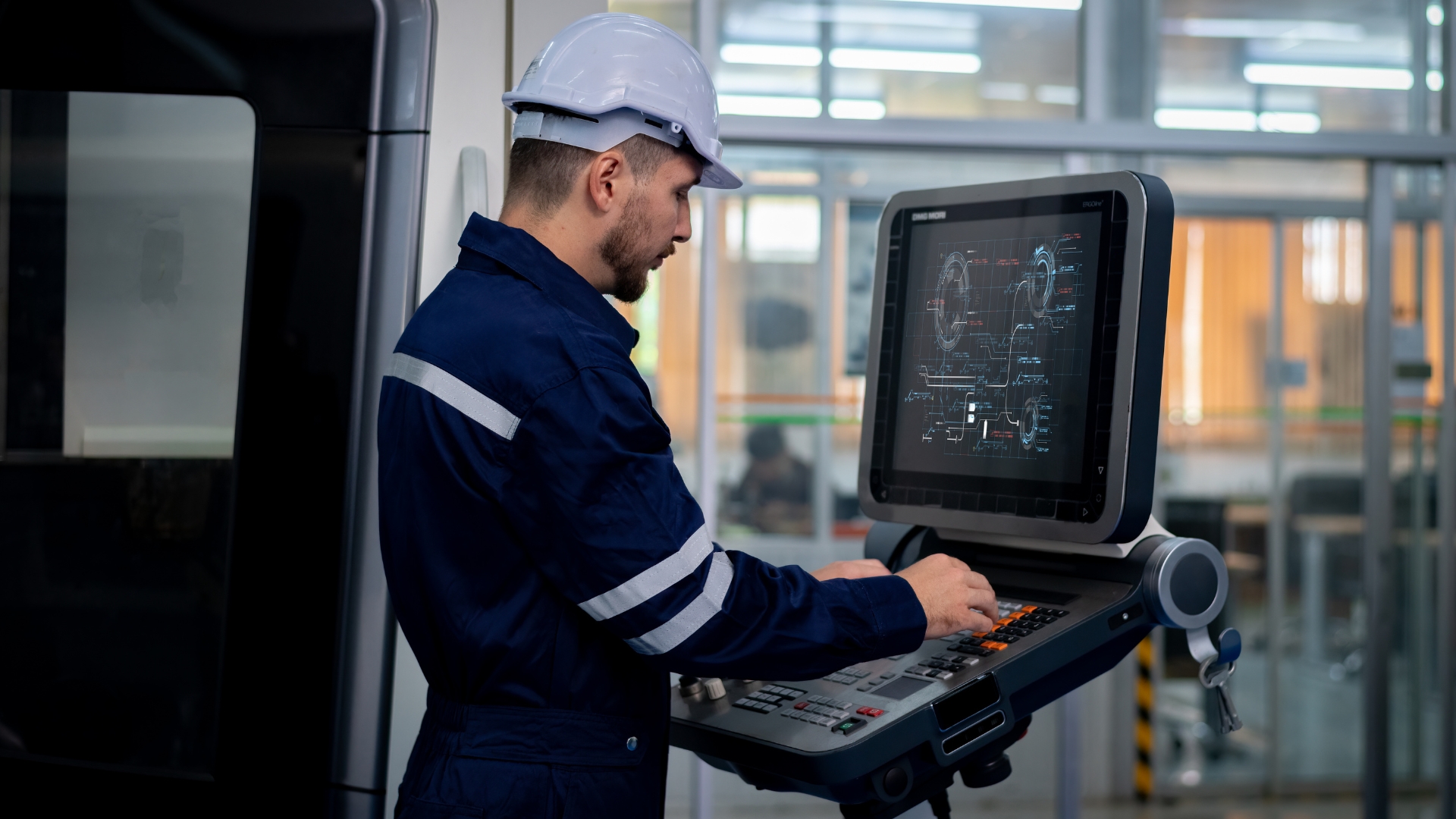 A CNC machinist wearing a white hard hat and blue work uniform operating a computerized control panel in a precision manufacturing facility.